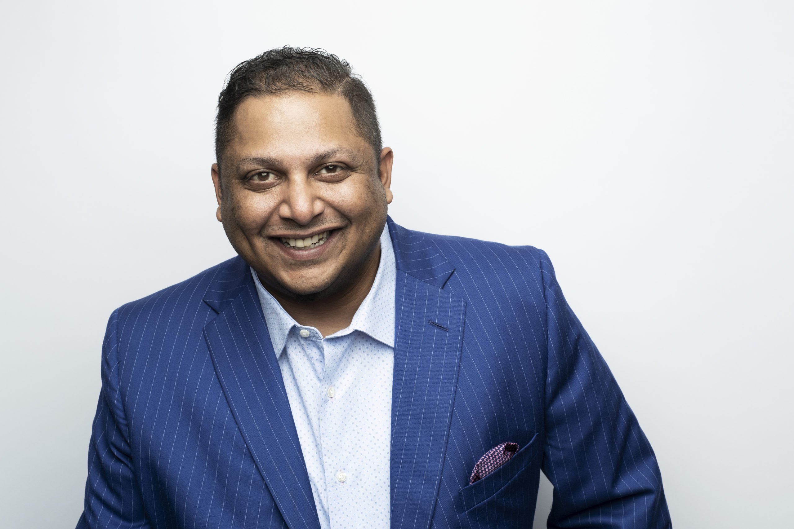 Vikash Patel A man with short dark hair, wearing a blue pinstripe suit and a light blue shirt, smiling broadly in front of a plain white background.