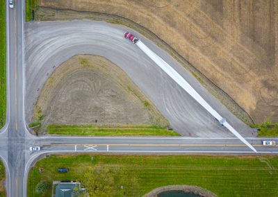 Aerial view of a large truck transporting a long wind turbine blade along a curved gravel road intersecting farmland and paved roads, with cars and fields visible nearby.