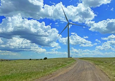 A large wind turbine stands in a grassy field next to a winding dirt road under a bright blue sky filled with fluffy white clouds.