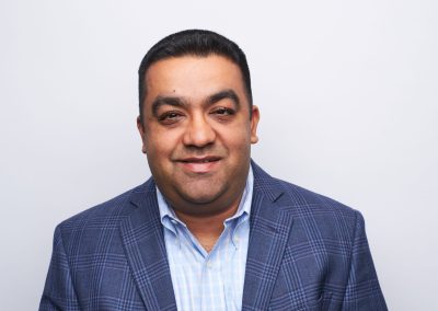 A man with short dark hair, wearing a blue plaid blazer and a light blue collared shirt, smiles slightly while facing the camera against a plain white background.
