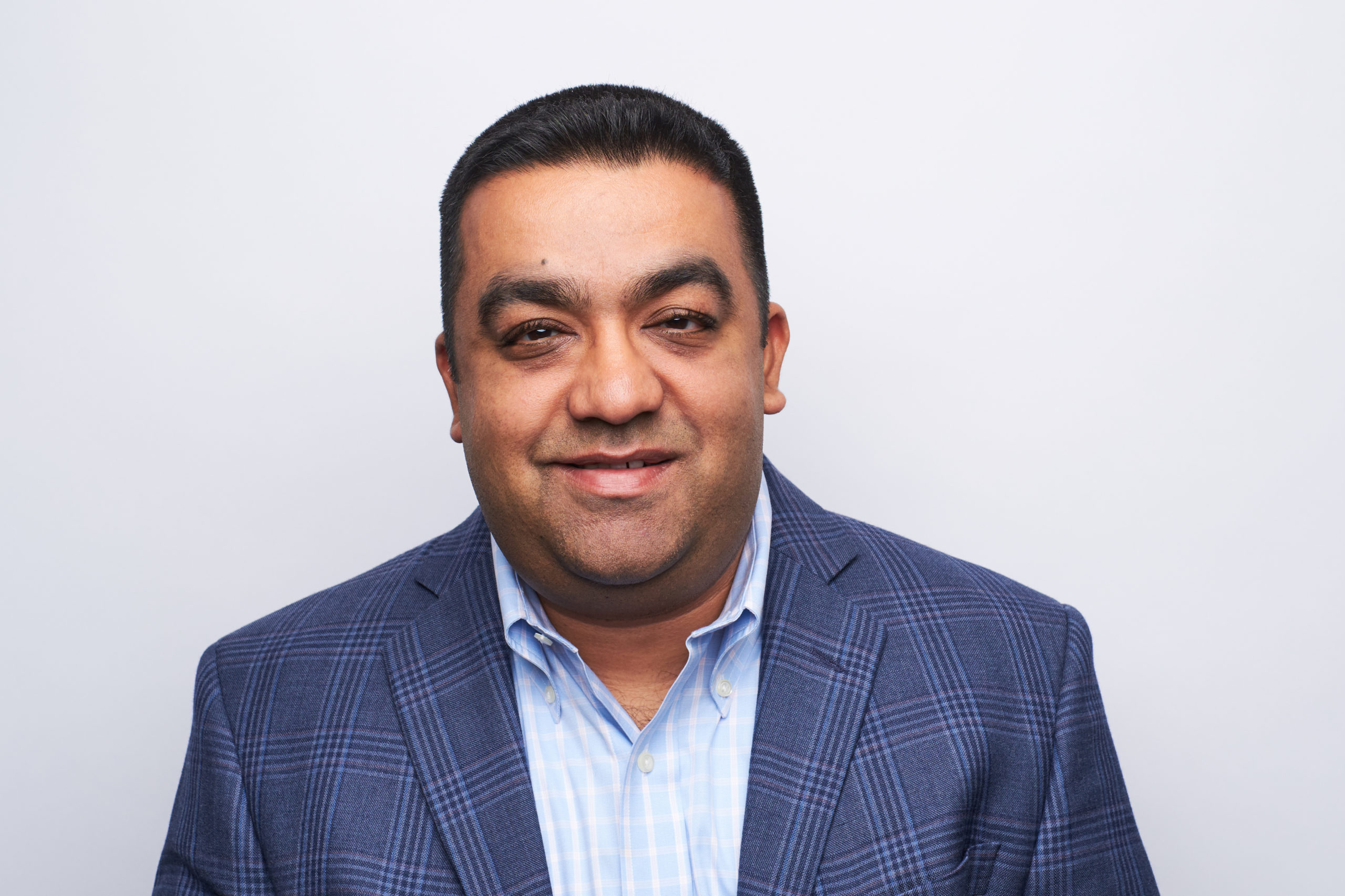 Mihir Patel A man with short dark hair, wearing a blue plaid blazer and a light blue collared shirt, smiles slightly while facing the camera against a plain white background.