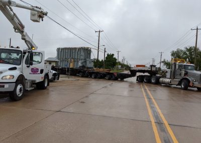 A large truck hauling an oversized load blocks both lanes of a wet road, while a utility vehicle with a raised boom is parked nearby under cloudy skies.