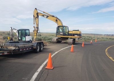Excavators and a truck are parked on the side of a curved rural road, with orange traffic cones marking the work zone. The sky is partly cloudy, and the surrounding landscape is open and grassy.