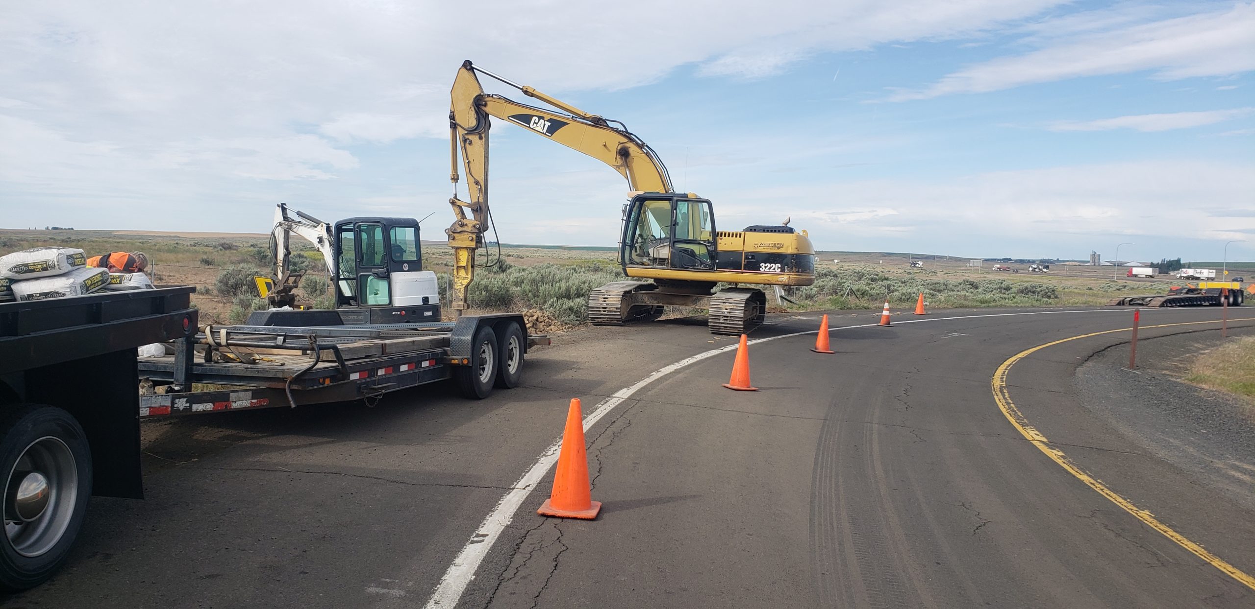 Excavators and a truck are parked on the side of a curved rural road, with orange traffic cones marking the work zone. The sky is partly cloudy, and the surrounding landscape is open and grassy.