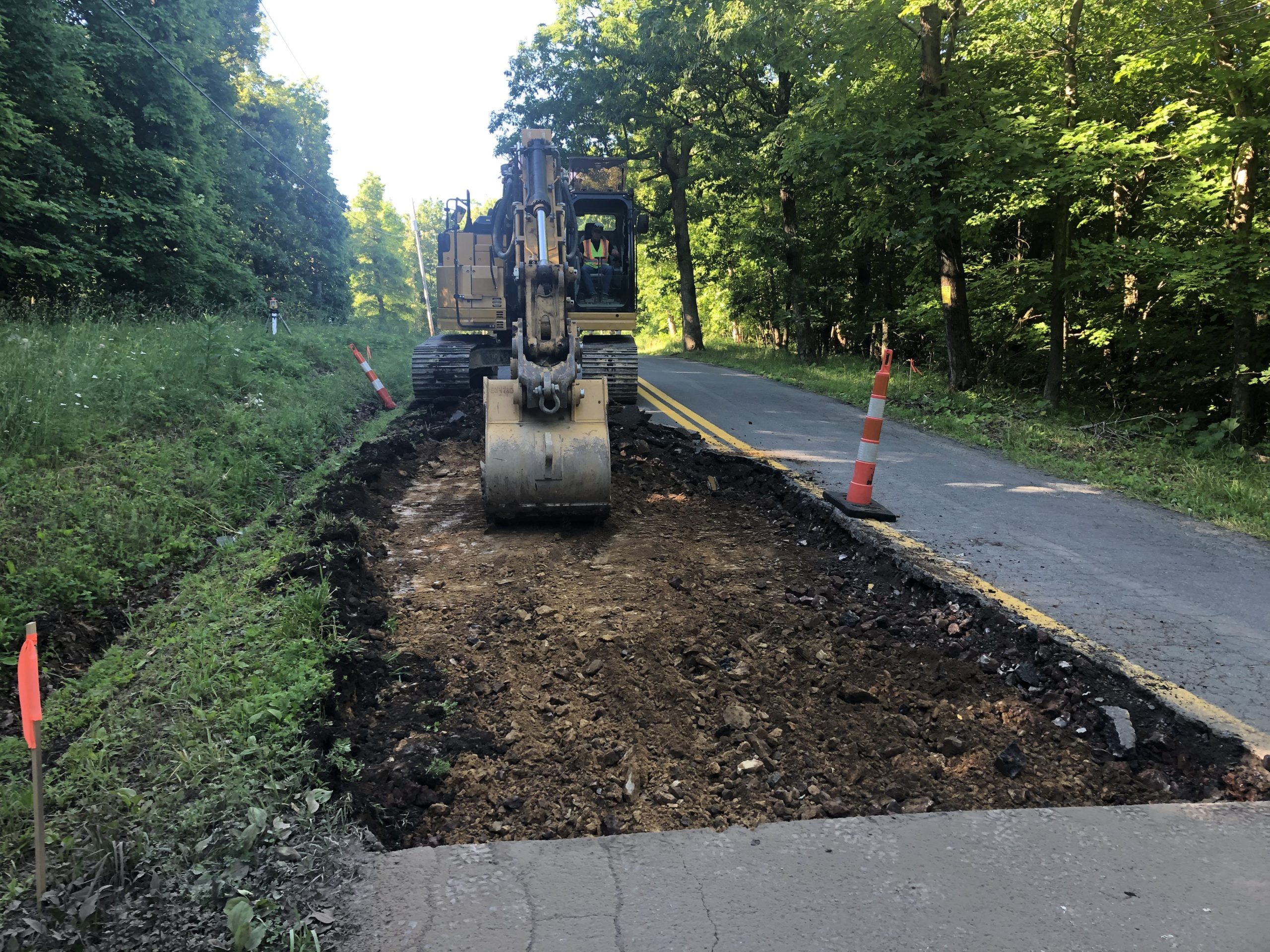 7:19 post A construction vehicle is digging up a section of a rural road, with traffic cones and flags marking the area. Trees and greenery line both sides of the road under bright daylight.