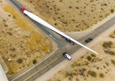Aerial view of a long truck transporting a single, massive wind turbine blade on a desert road, escorted by two smaller vehicles at an intersection.