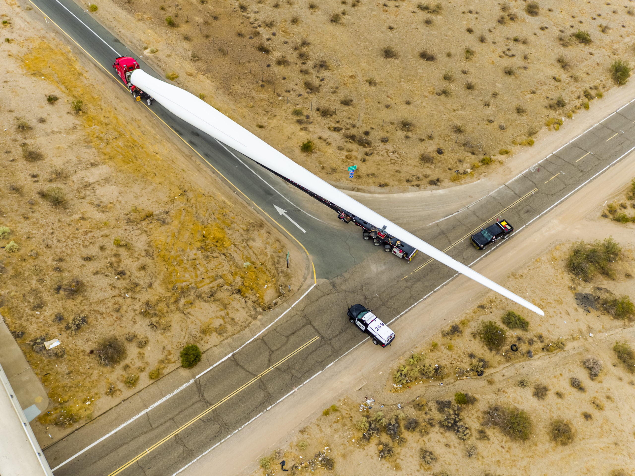Golden State Aerial view of a long truck transporting a single, massive wind turbine blade on a desert road, escorted by two smaller vehicles at an intersection.