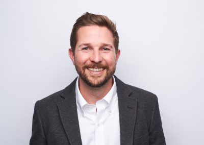 A man with short brown hair and a beard smiles at the camera. He is wearing a white shirt and a dark gray blazer, standing against a plain white background.