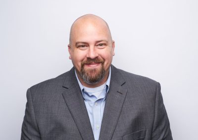 A smiling bald man with a goatee wearing a gray suit jacket and light blue shirt, posing against a plain white background.
