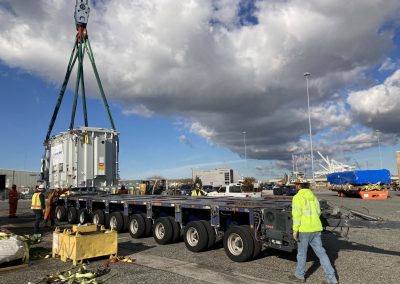Workers in safety gear guide a large piece of industrial equipment being hoisted by a crane onto a multi-axle flatbed trailer at a busy outdoor industrial site under a partly cloudy sky.
