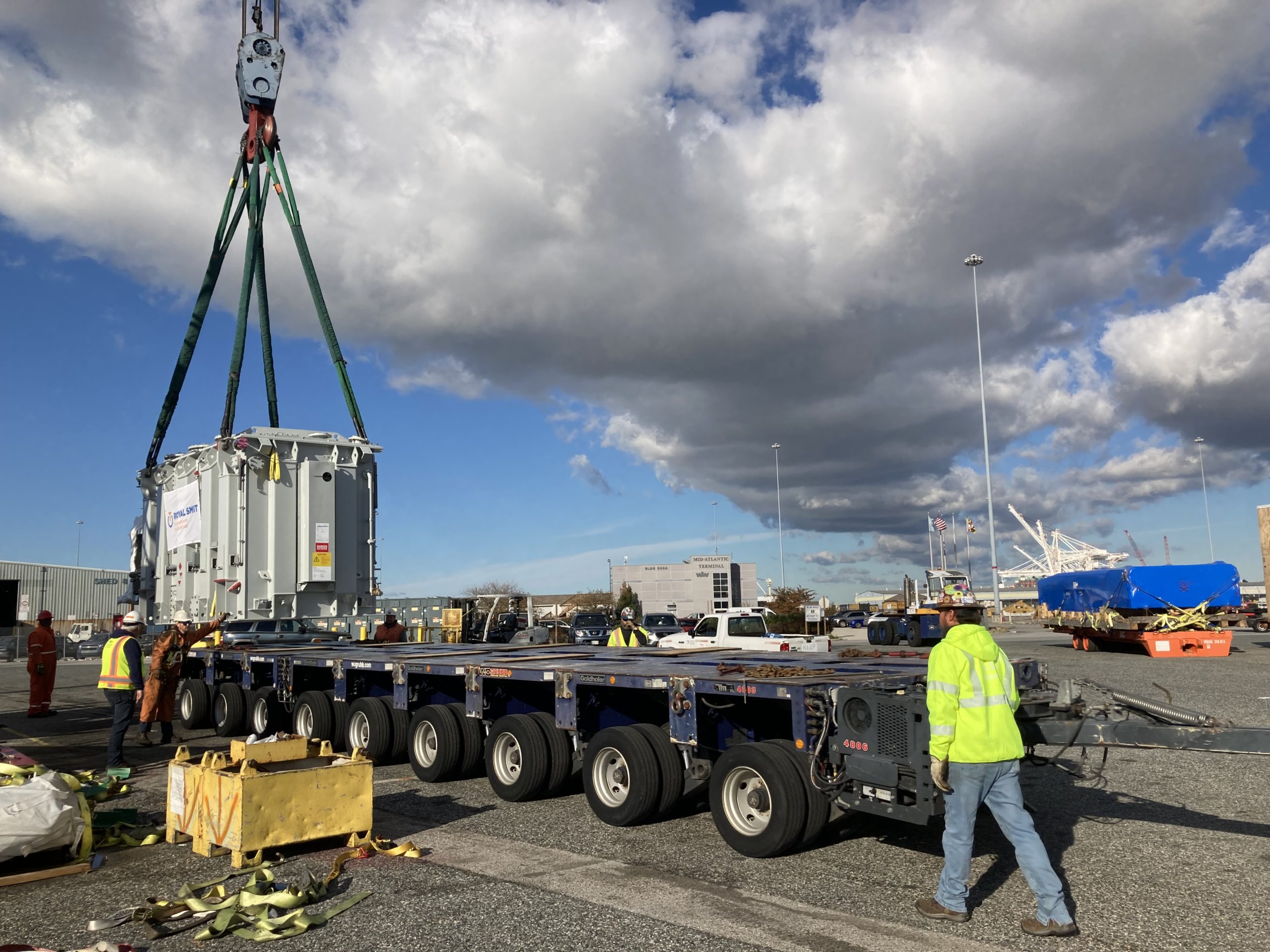 IMG_1720 Workers in safety gear guide a large piece of industrial equipment being hoisted by a crane onto a multi-axle flatbed trailer at a busy outdoor industrial site under a partly cloudy sky.