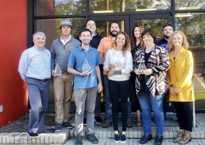A group of ten people, some holding glass awards, stand and smile outside a building with large windows and a red wall. They appear to be celebrating an achievement or recognition.