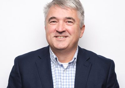 Smiling middle-aged man with short gray hair, wearing a navy blazer over a light blue and white checkered shirt, standing in front of a plain white background.
