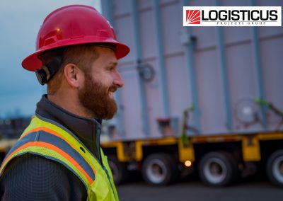 A bearded man in a red hard hat and safety vest smiles while standing outdoors near industrial equipment. The LOGISTICUS Projects Group logo appears in the top right corner.
