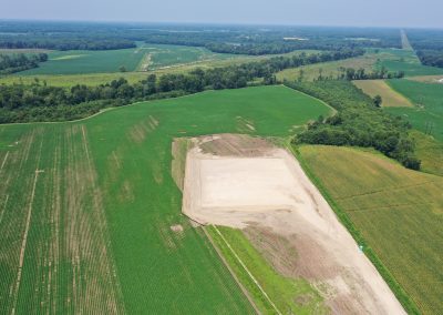 Aerial view of green farmland with a large rectangular cleared dirt area in the center, surrounded by fields and patches of trees under a clear sky.