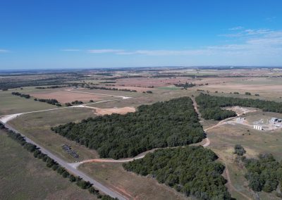 Aerial view of a rural landscape with open fields, patches of dense trees, winding dirt roads, and a few scattered buildings under a clear blue sky.