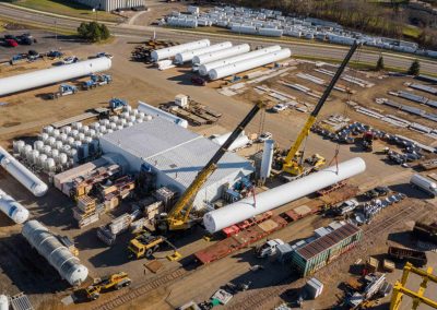 Aerial view of an industrial facility with cranes lifting large cylindrical tanks, surrounded by numerous white storage tanks, machinery, trucks, and various equipment arranged on the ground.
