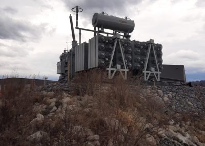 A large electrical transformer sits on rocky ground under a cloudy sky, surrounded by dry grass and stones, with some distant hills visible in the background.