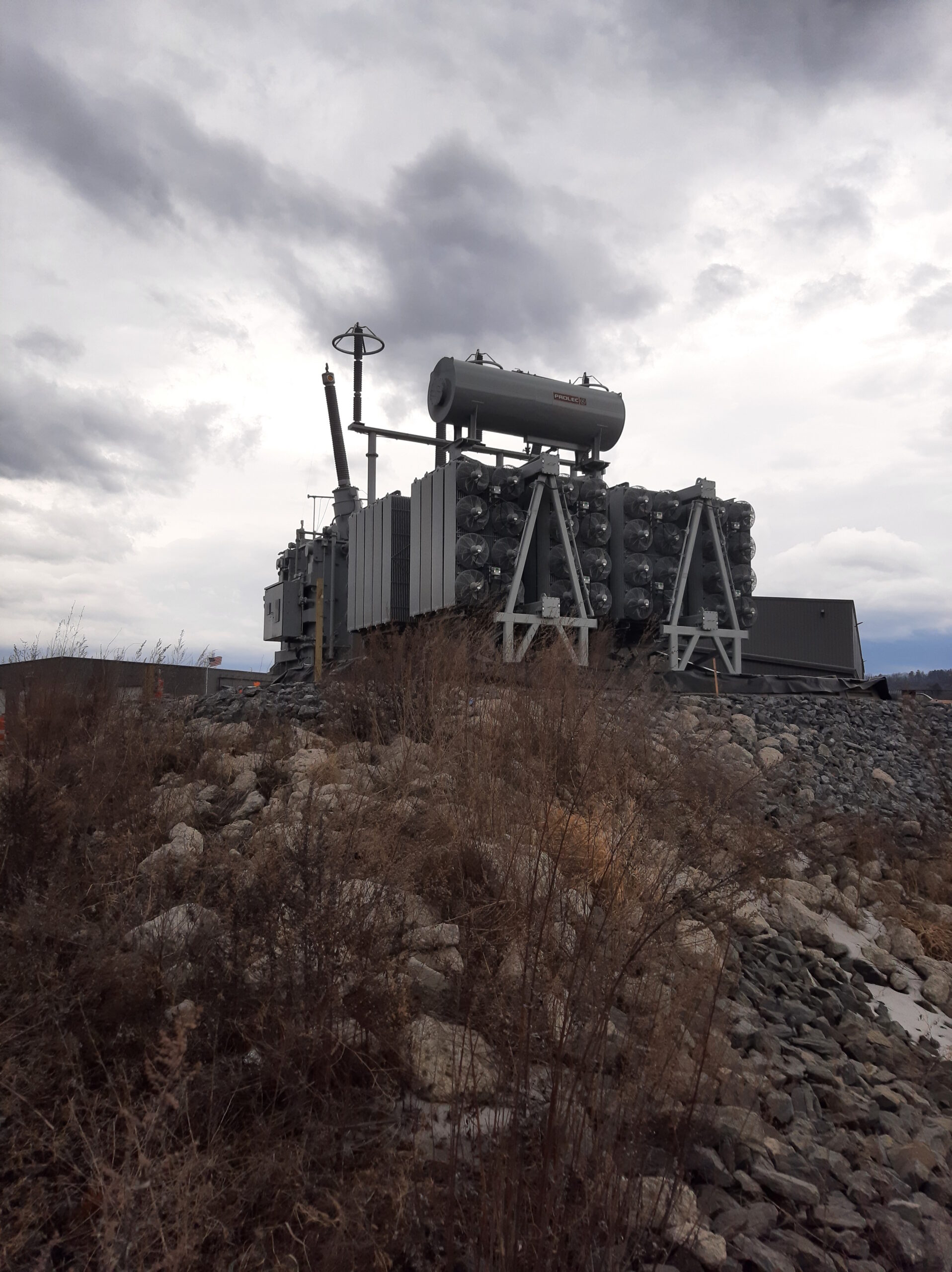 LG A large electrical transformer sits on rocky ground under a cloudy sky, surrounded by dry grass and stones, with some distant hills visible in the background.