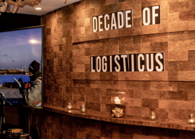 A textured brown wall displays the words DECADE OF LOGISTICS with some letters misaligned. To the left, a large screen shows an outdoor scene with vehicles and a person in safety gear near a body of water. Candles sit on a narrow shelf.