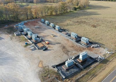Aerial view of a construction site with several rectangular building foundations, stacks of construction materials, and machinery, surrounded by grassy fields and trees in the background.