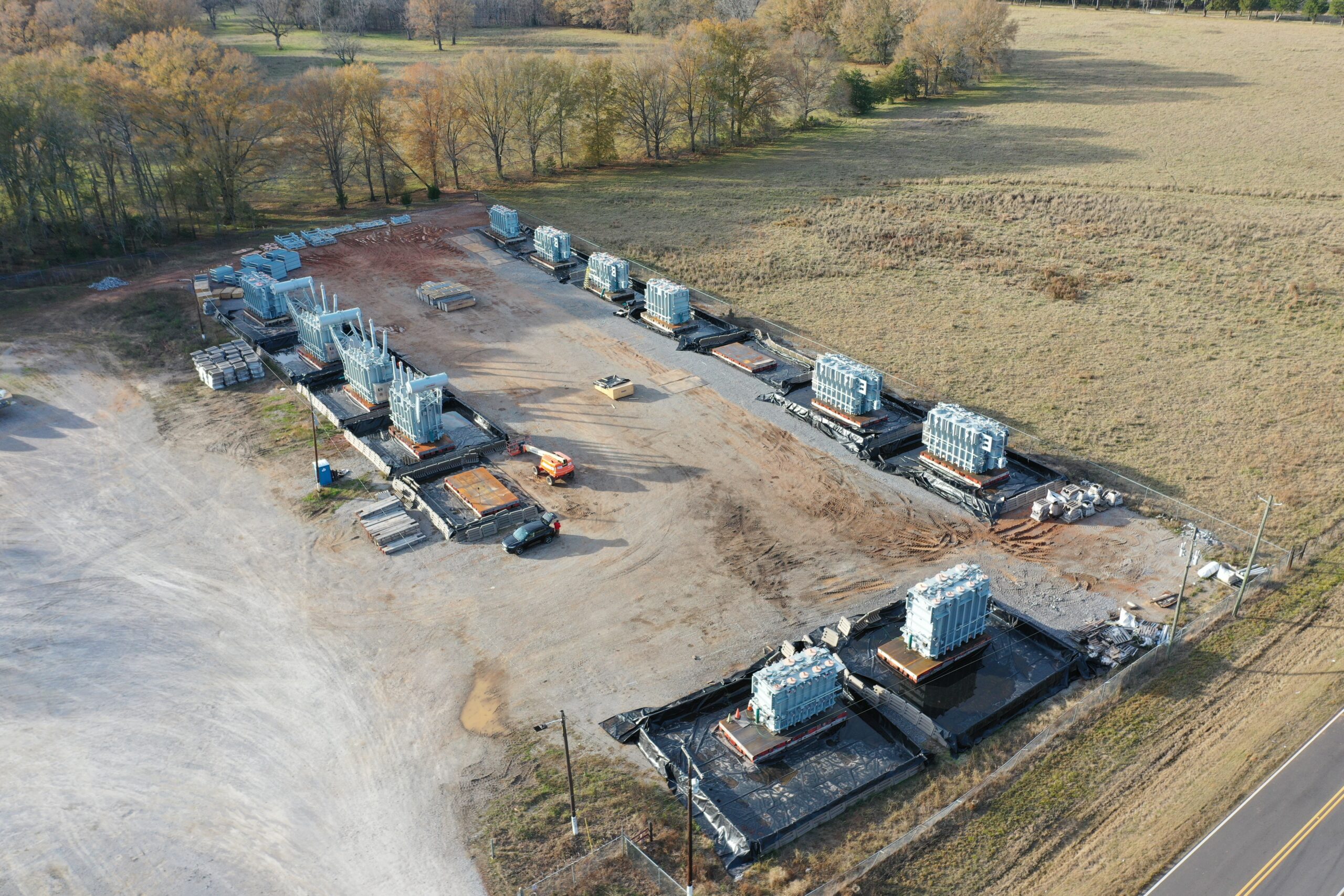 default Aerial view of a construction site with several rectangular building foundations, stacks of construction materials, and machinery, surrounded by grassy fields and trees in the background.