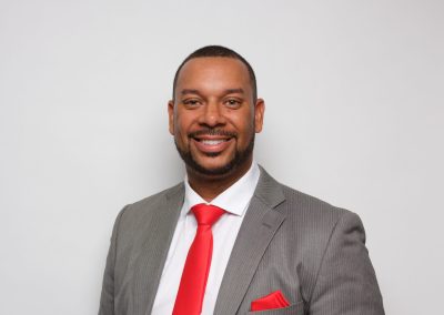 A man in a gray suit with a red tie and matching pocket square smiles at the camera against a plain white background.