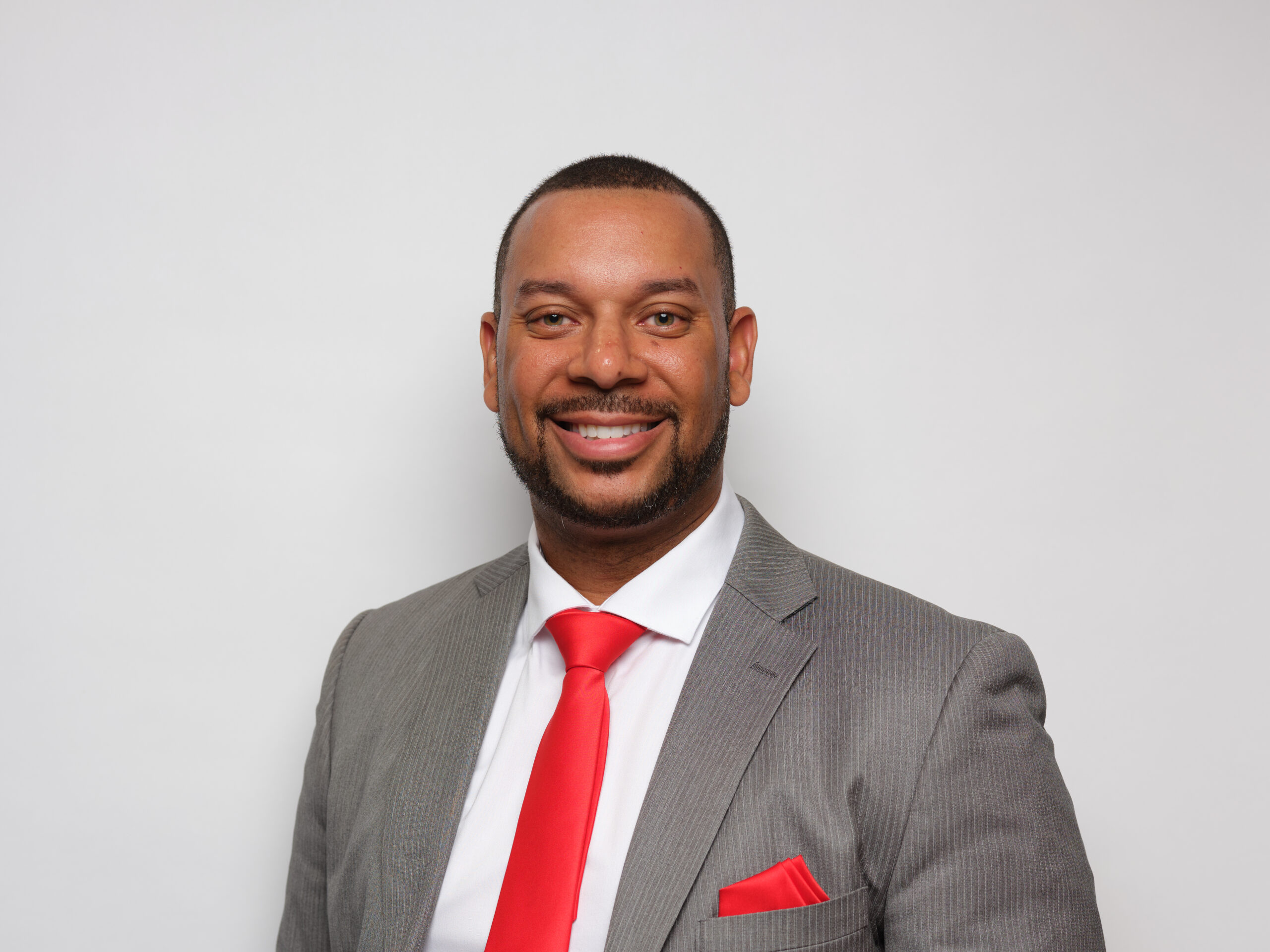 A man in a gray suit with a red tie and matching pocket square smiles at the camera against a plain white background.