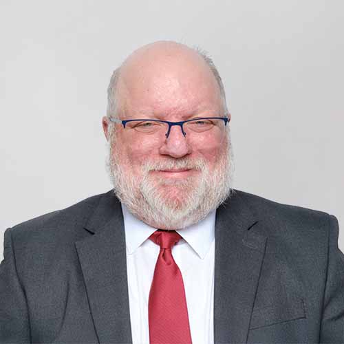 Smiling Chris King with a white beard, glasses, and a bald head is wearing a dark suit, white shirt, and red tie, posing against a plain light gray background.