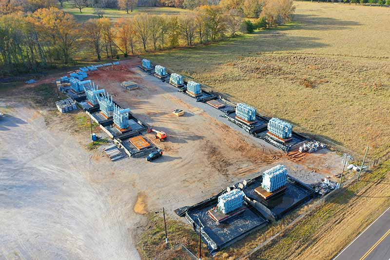 Aerial view of a construction site with multiple building foundations and stacks of materials, surrounded by grassy fields and trees, near a road on the right side of the image.