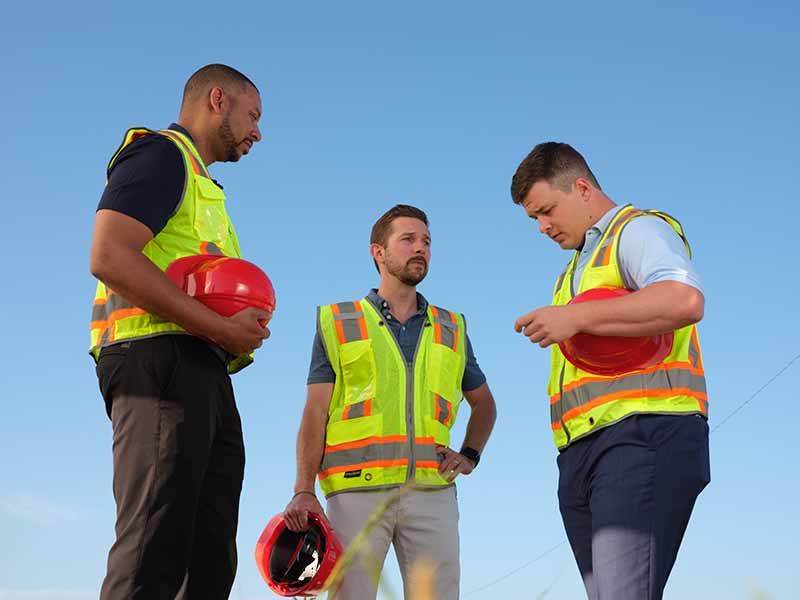 Three men wearing safety vests and holding red hard hats stand outdoors under a clear blue sky, appearing to discuss something work-related.