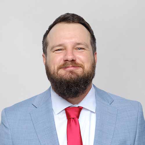 Dariusz Stochawiak with a brown beard and short hair smiles at the camera. He is wearing a light blue suit jacket, white shirt, and red tie, posed against a plain light gray background.