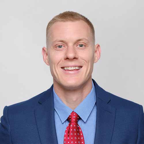 Devin Secore with short blond hair, wearing a blue suit, light blue shirt, and red patterned tie, smiles at the camera against a plain light background.