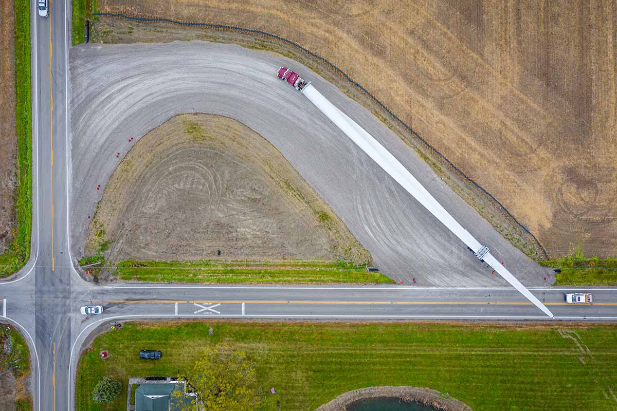 Aerial view of a long truck transporting a wind turbine blade, turning at a rural intersection surrounded by fields, grass, and a few cars on the road.