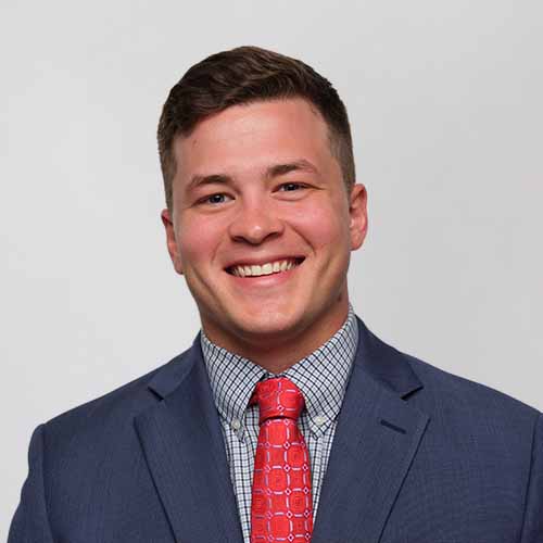 Smiling Jackson Clark in a blue suit, patterned shirt, and red patterned tie stands against a plain light background.