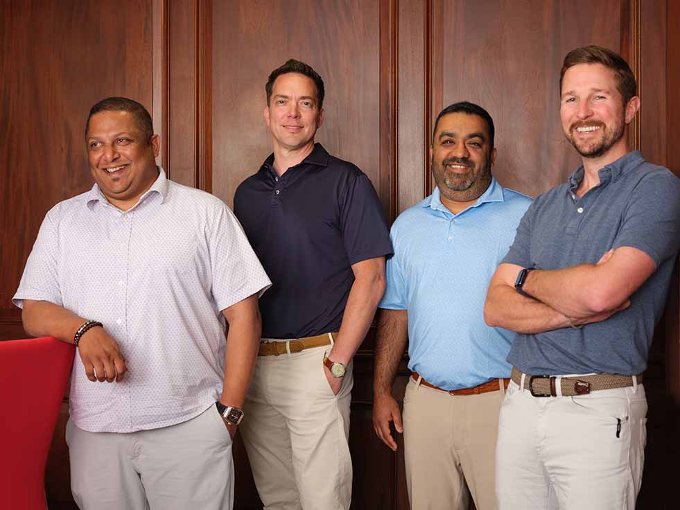 Logisticus Leadership Team stand in front of a wooden panel wall, smiling at the camera. They are dressed in casual, collared shirts and light-colored pants. One man leans on a red chair.