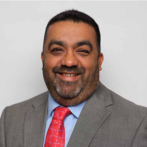 Mihir Patel with short dark hair and a beard, wearing a grey suit, blue shirt, and red patterned tie, smiling in front of a plain light background.