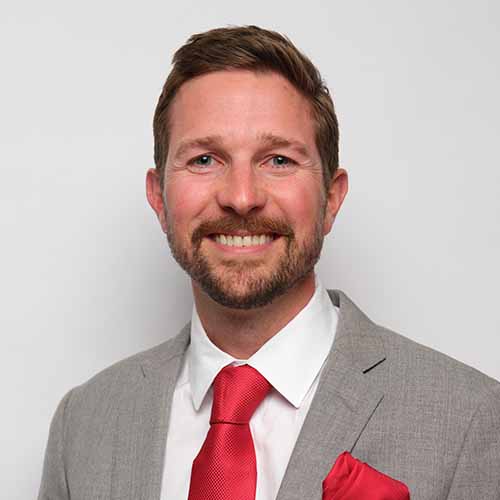 Smiling Phillip Mitchum with short brown hair and beard wearing a light gray suit, white shirt, and bright red tie with a matching red pocket square, standing in front of a plain white background.