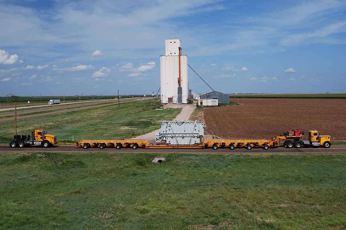 A large yellow truck convoy transports an oversized load on a rural road, passing farmland with a tall white grain elevator and blue sky in the background.