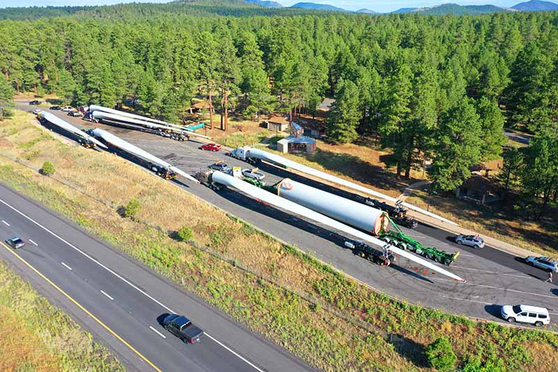 Aerial view of several large wind turbine blades loaded on trucks parked at a rest area near a highway, surrounded by dense green forest and a few small buildings.