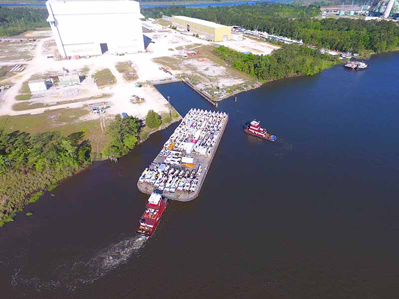 Aerial view of a large barge loaded with cargo, escorted by two red tugboats on a river near a shoreline with trees and an industrial facility in the background.