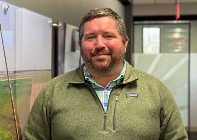A man with short brown hair and a beard, wearing a green Patagonia fleece over a blue plaid shirt, stands indoors in an office hallway with artwork and frosted glass panels in the background.