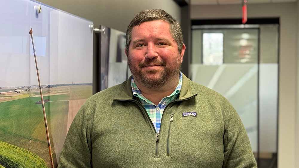 A man with short brown hair and a beard, wearing a green Patagonia fleece over a blue plaid shirt, stands indoors in an office hallway with artwork and frosted glass panels in the background.