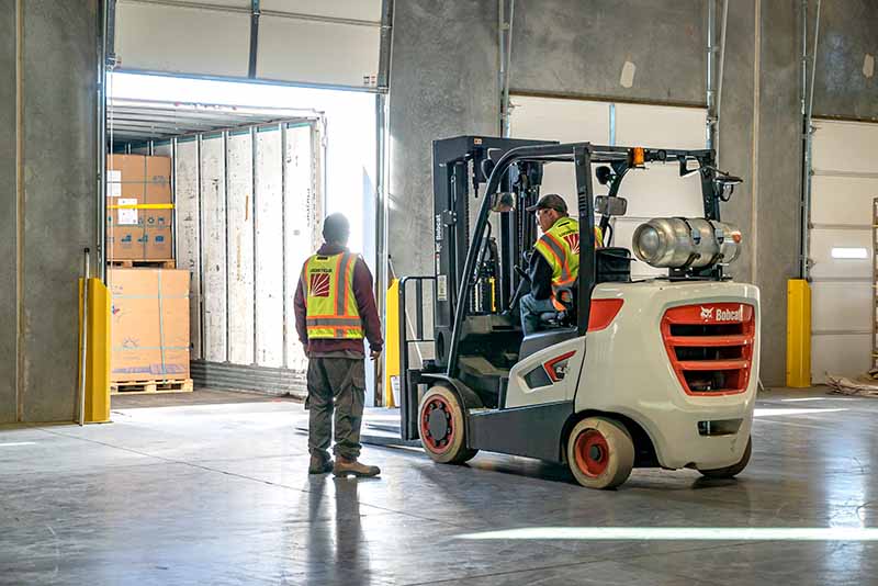 Two workers wearing safety vests load boxes into a truck at a warehouse; one operates a forklift while the other stands nearby, overseeing the loading process.