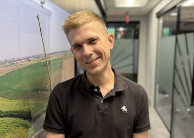 A smiling man with short blonde hair wearing a black polo shirt stands in a modern office hallway, next to a wall with a large photo of a green landscape.