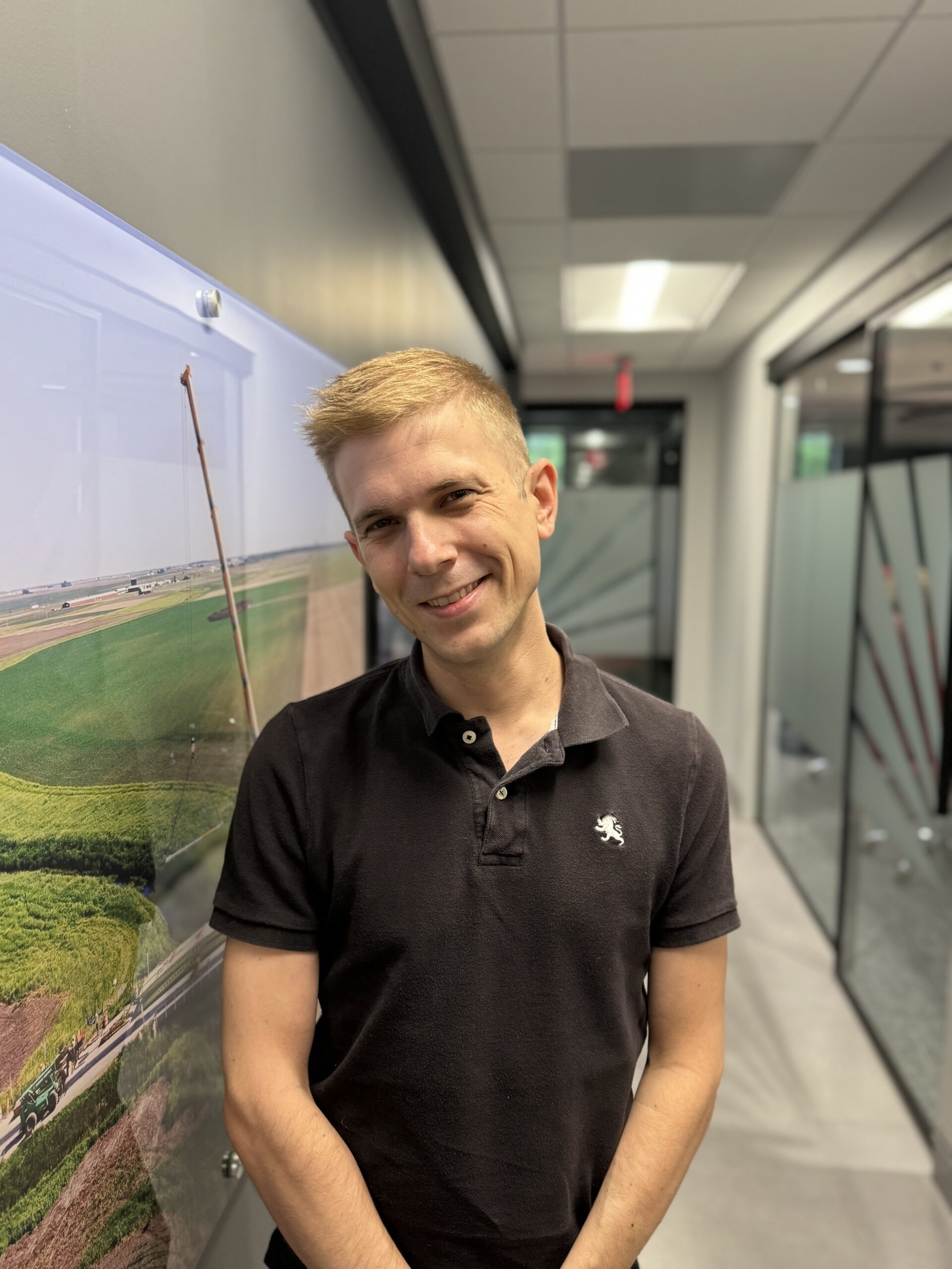 A smiling man with short blonde hair wearing a black polo shirt stands in a modern office hallway, next to a wall with a large photo of a green landscape.