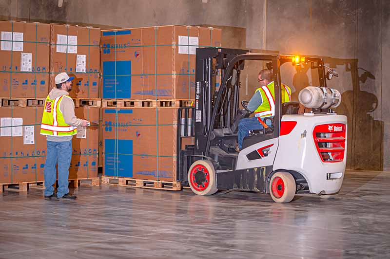 A warehouse worker operates a forklift, moving large stacked boxes while another worker in a safety vest and hard hat stands nearby, guiding the process.