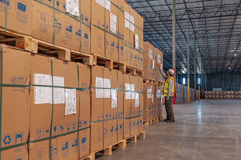 A worker in a safety vest and helmet stands in a large warehouse, looking at tall stacks of large cardboard boxes on wooden pallets arranged in multiple rows.