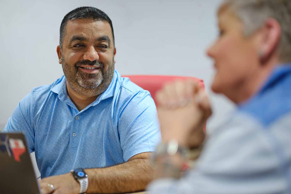 A man in a blue shirt smiles while talking to another person, who is out of focus in the foreground. They appear to be having a friendly conversation at a desk.