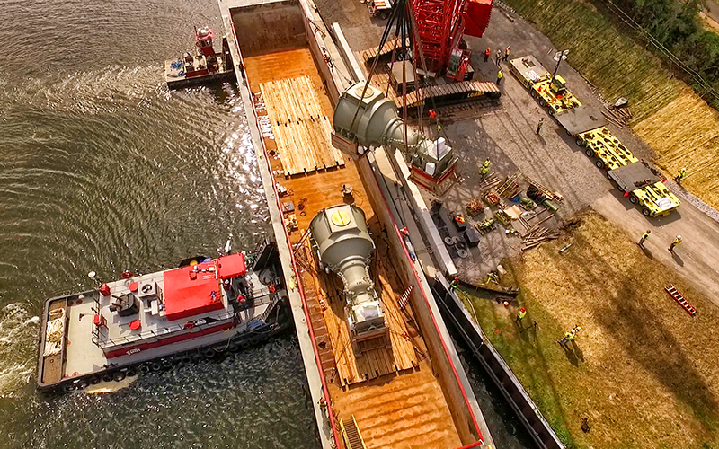 multi mode transport page Aerial view of a barge at a riverside construction site, with a crane lifting large industrial equipment onto the vessel; workers and vehicles are positioned along the shore and dock.